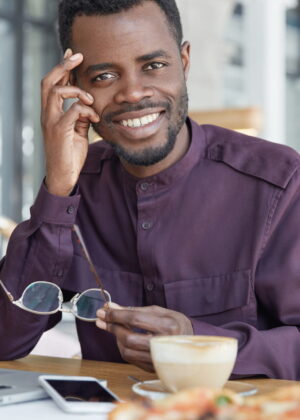 Happy dark skinned male freelancer uses modern electronic gadgets for remote work, sits against cozy coffee shop interior, drinks aromatic espresso, thinks about new successful approaches in business